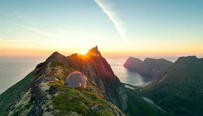 Grønt fjell står på en fjellrygg som leder mot et spisst fjell. I bakgrunnen går sola ned i havet.