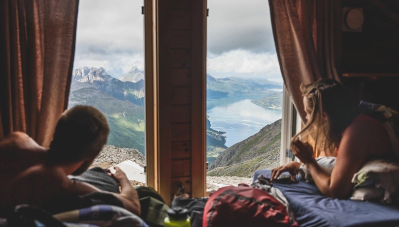 To personer ligger i senger på hver side av et vindu og skuer utover fjord og fjell.