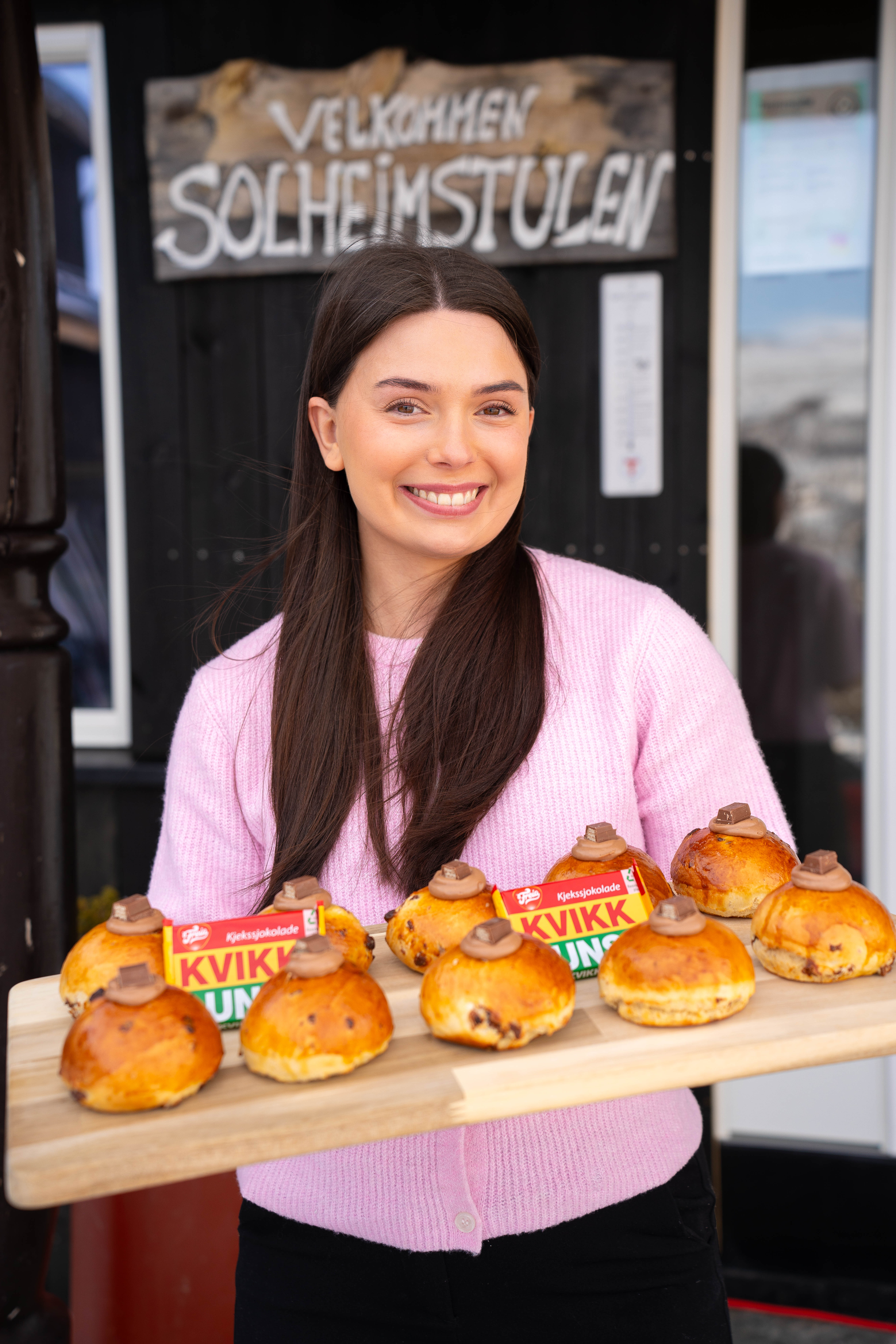 en kvinne med brunt h&aring;r og rosa og hvit bluse, smiler til kameraet mens hun holder en trefj&oslash;l med boller og bokser med solo. 