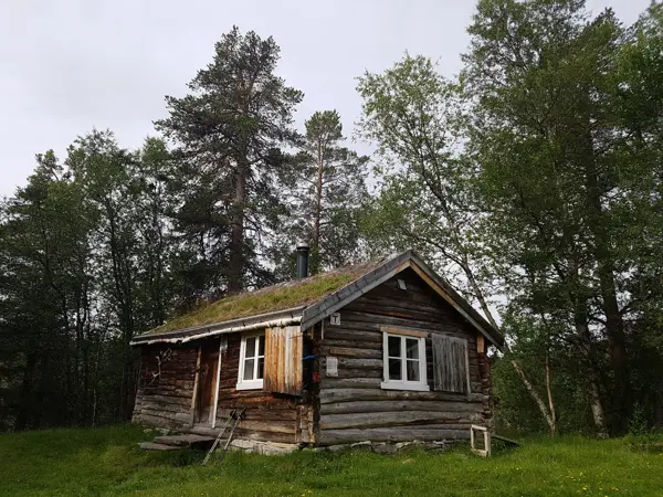 Ei gråbrun hytte på ei gresslette. Høye trær bak.