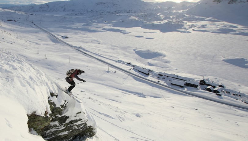 Skikjører hopper ned en liten klippe i en bratt fjellside ovenfor ei turisthytte.