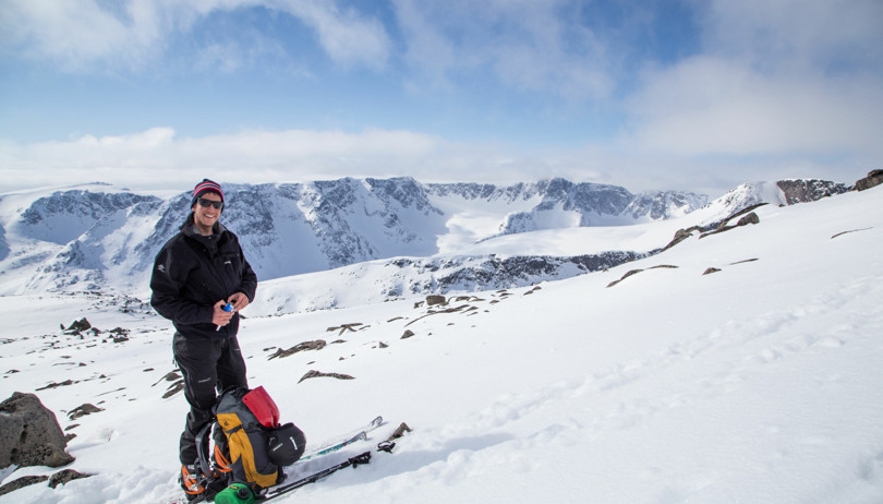 Skiløper med sekk og staver på bakken foran seg. Høye og bratte fjell i bakgrunnen.