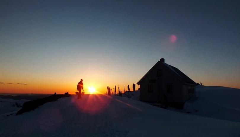 Silhuett av en person som går mot ei hytte i solnedgang.