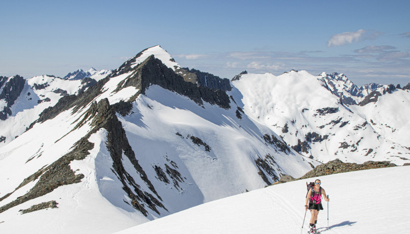 Kvinne i rosa singlet og svart skjørt går på ski opp ei fjellside i alpint landskap.