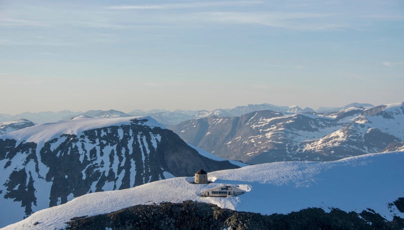 Et steintårn og ei hytte toppen av et snødekt fjell. I bakgrunnen skimtes fjell og daler.