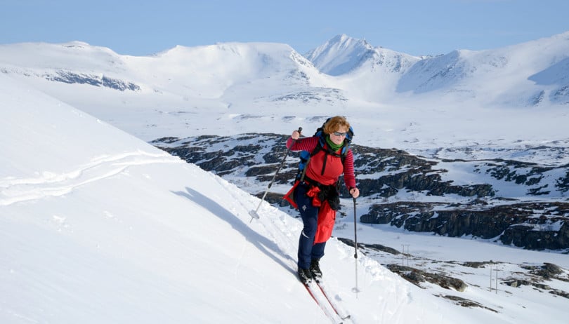Kvinne i rød genser og bukse kjører ned en liten bakke på ski. I bakgrunnen skimtes høye og snøkledde fjell.