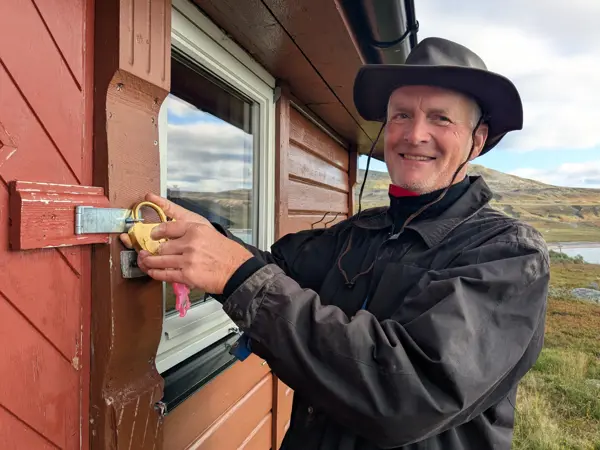 En mann med sorte fjellklær og hatt låser opp en hengelås på døra til ei rødbrun fjellhytte. Man skimter et vann i bakgrunnen.