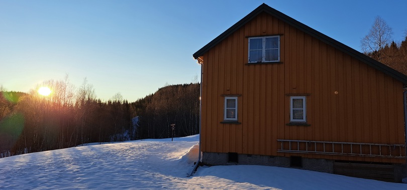 Ei gul hytte, vinter og snø. Skog og fjell i bakgrunnen. Sola skinner såvidt over tretoppene.