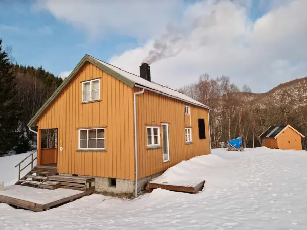 Ei gul hytta med et gult lite uthus bak. Det er snø på hytta. Røyk fra pipa. Granskog i bakgrunnen.