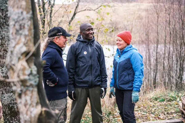 Tre personer står og samtaler i et skogsholdt, høstlige farger på naturen rundt.