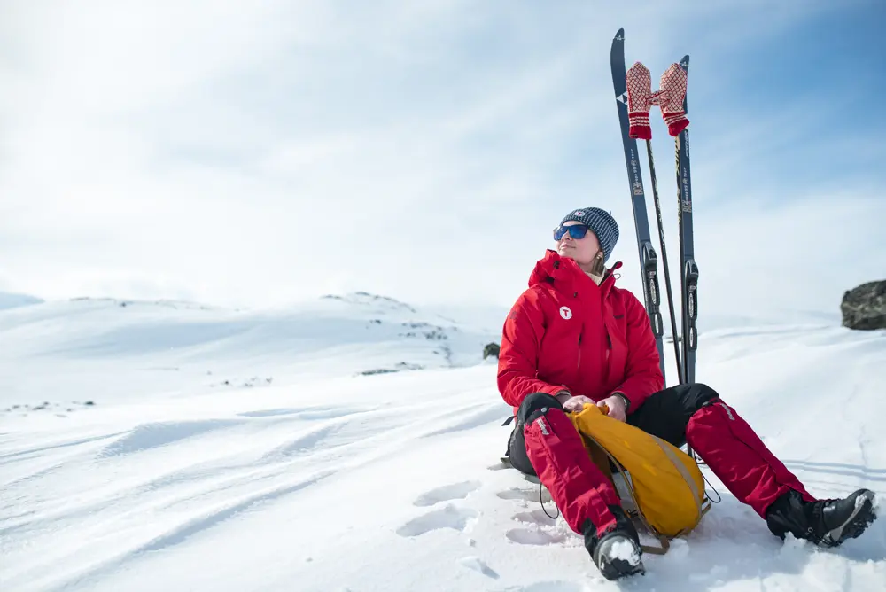 Kvinne i rød jakke sitter i snøen. Hun ser fornøyd ut. Ved siden av henne ses ski og en oransje tur sekk