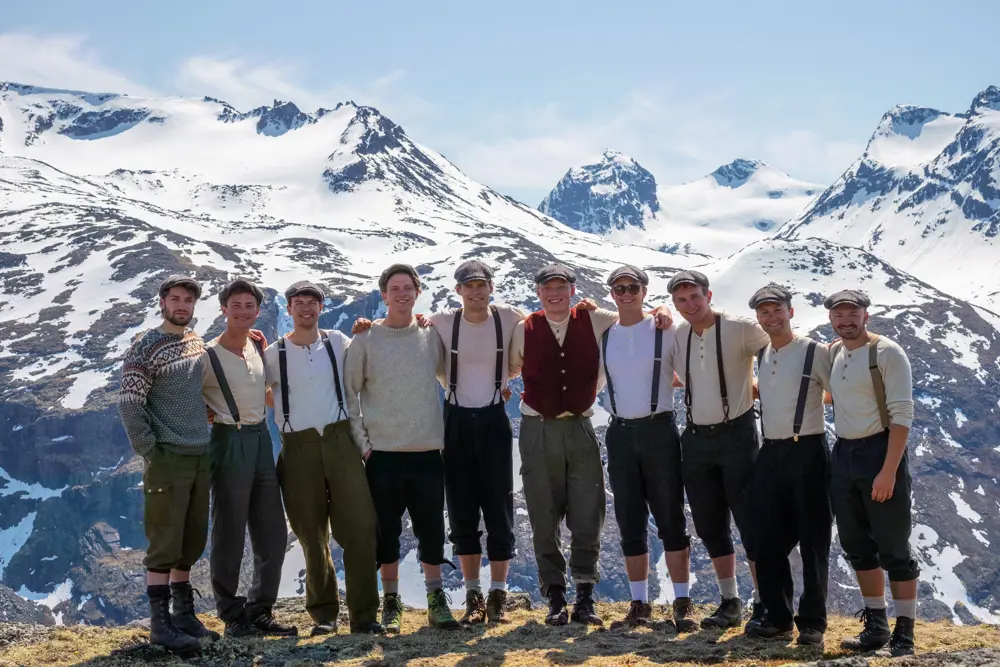 En rekke menn som står holder rundt hverandre og smiler til kamera. Bak ser vi snødekte fjell