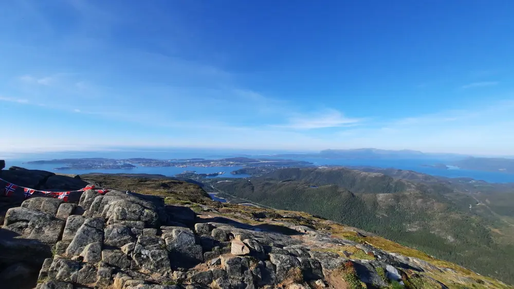 Bildet er tatt fra det som ser ut som en fjelltopp, med utsikt over skog og hav. Det er blå himmel.