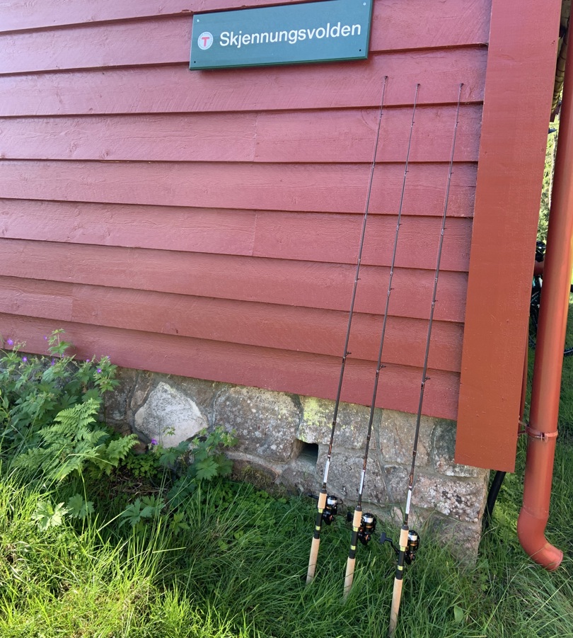 Tre fiskestenger som står lent inntil en rød hytte
