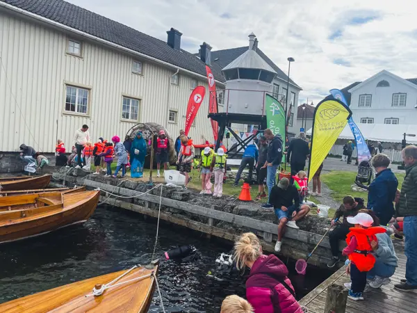 Barn og voksne samlet på en brygge, båter i sjøen og et fyr på brygga.