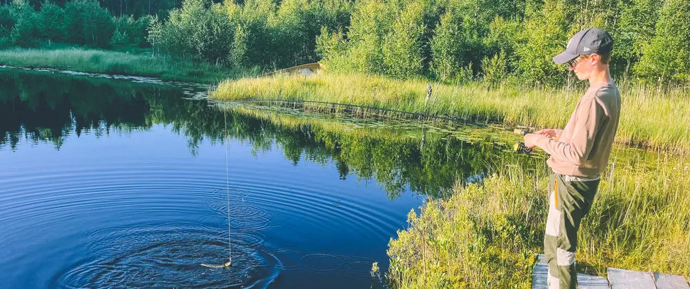Gutt fisker med stang i et skogstjern. Skog i bakgrunnen, og en skyfrihimmel speiler seg i vannet. 
