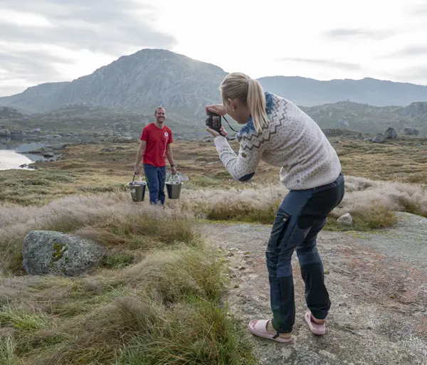 Kvinne i ullgenser fotograferer mann med rød t-skjorte som bærer to bøtter. De står i naturen