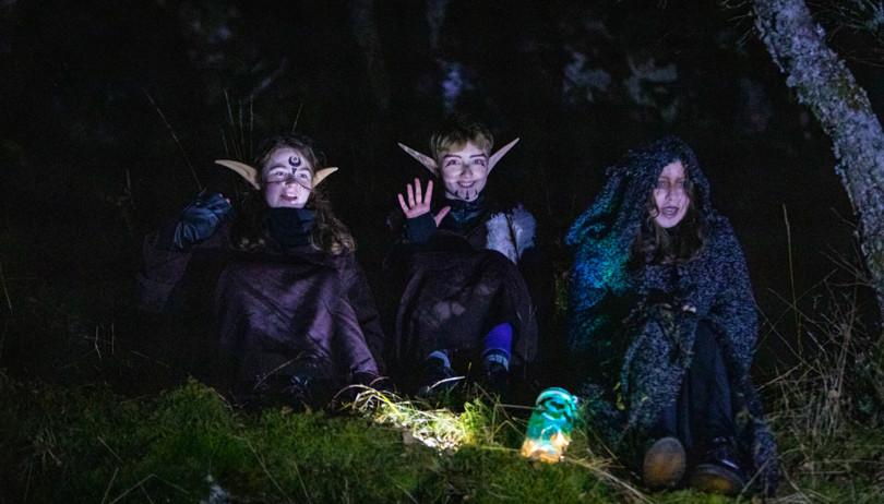 Tre barn utkledd i Halloween-kostyme sitter i mørket i gresset.