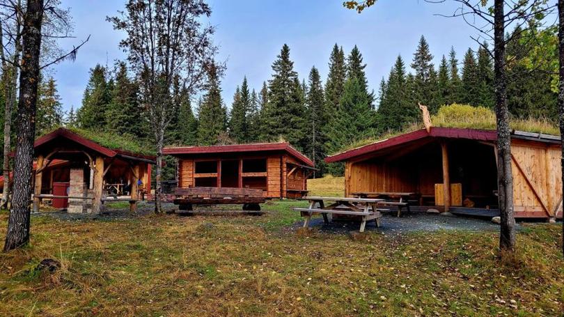 Bildet viser 3 gapahuker lager i tømmer med grass på taket, plassert på en stor lysning i skogen med flere sittebenker i midten.