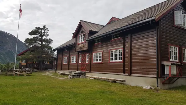 Brunbeiset hytte med stor furu og flaggstang utenfor, grønt gress på bakken og overskyet himmel.