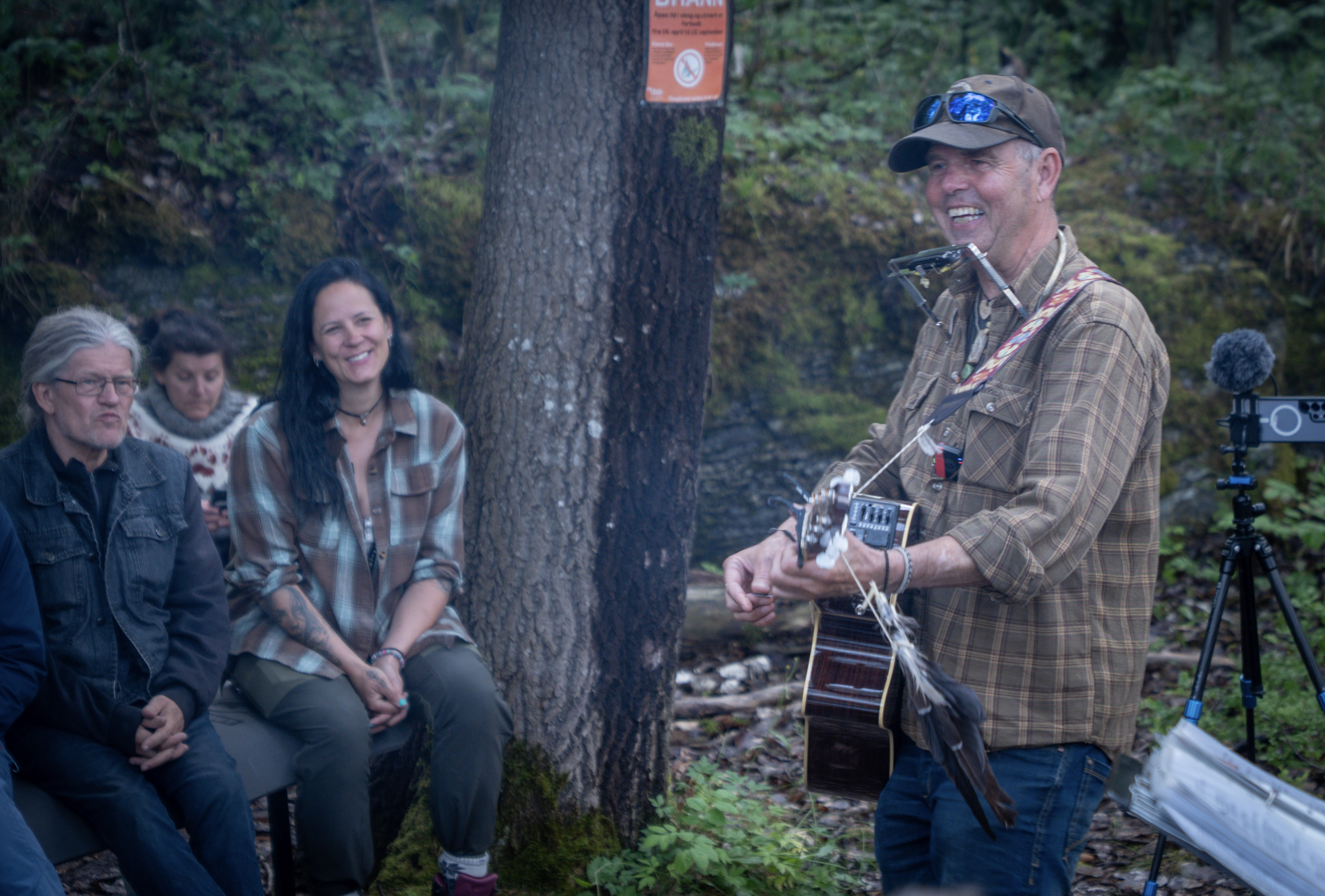 Smilende mann med gitar som har konsert ute i skogen. Foto.