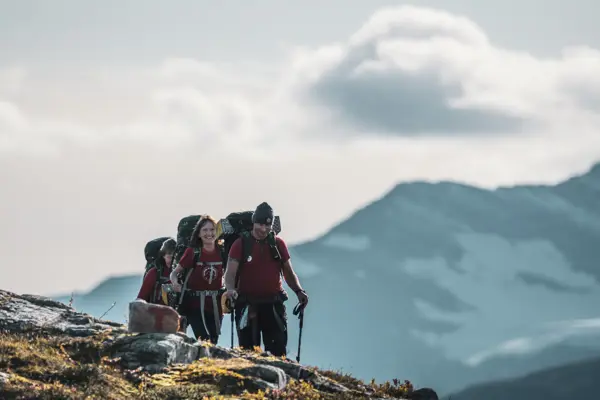 Tre mennesker med tursekker går i et fjellandskap. Et stort, snøkledt fjell i bakgrunnen. Alle går i T-skjorte, og mannen som går fremst har lue og gåstaver.