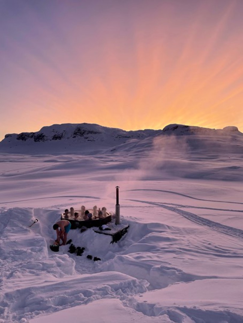 Flere mennesker i en varm stamp omkranset av et snørikt landskap med fjell og solnedgang i bakgunnen.