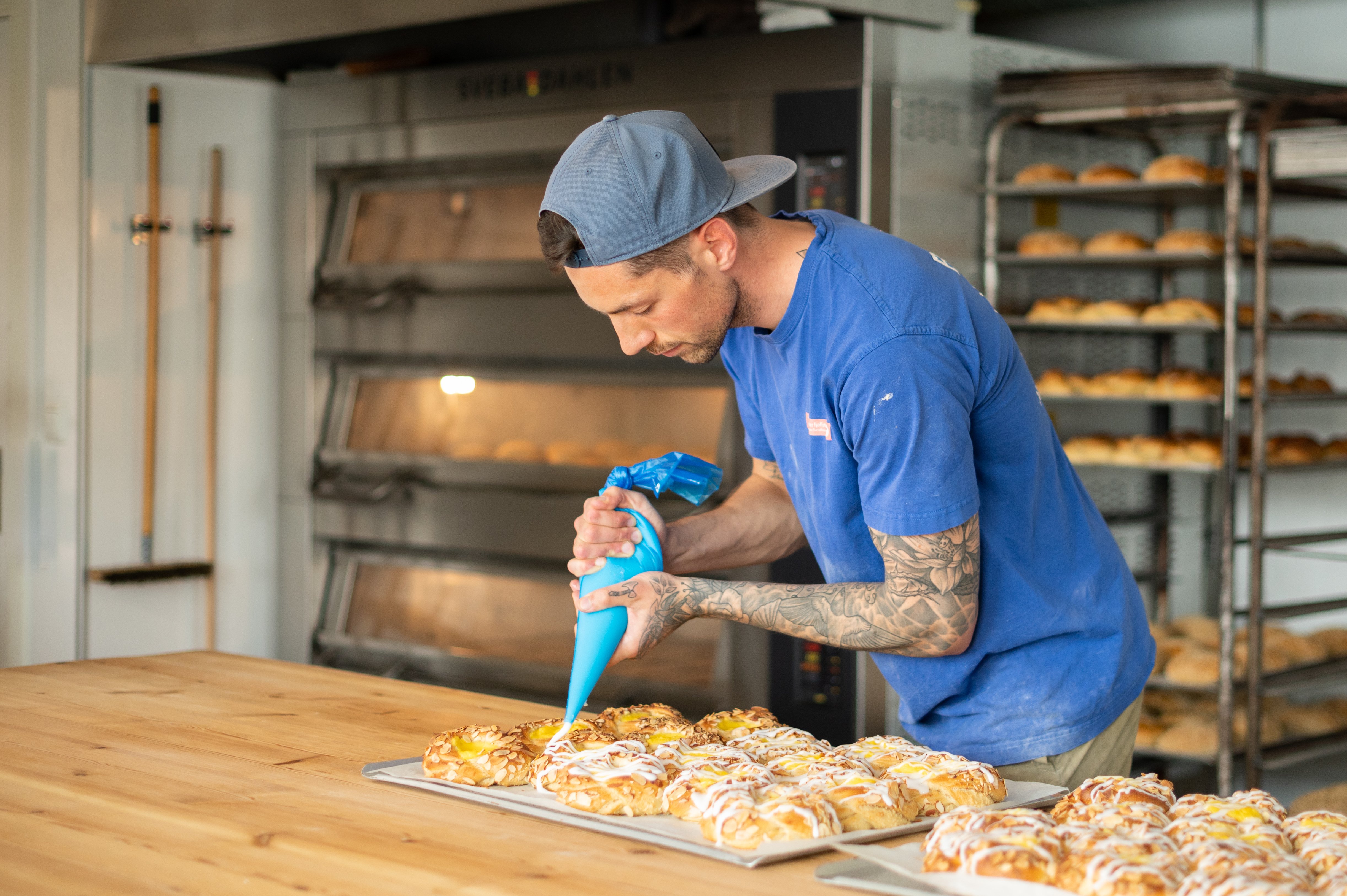 Foto. En mann st&aring;r i et bakeri og spr&oslash;yter glasur p&aring; et brett med boller.