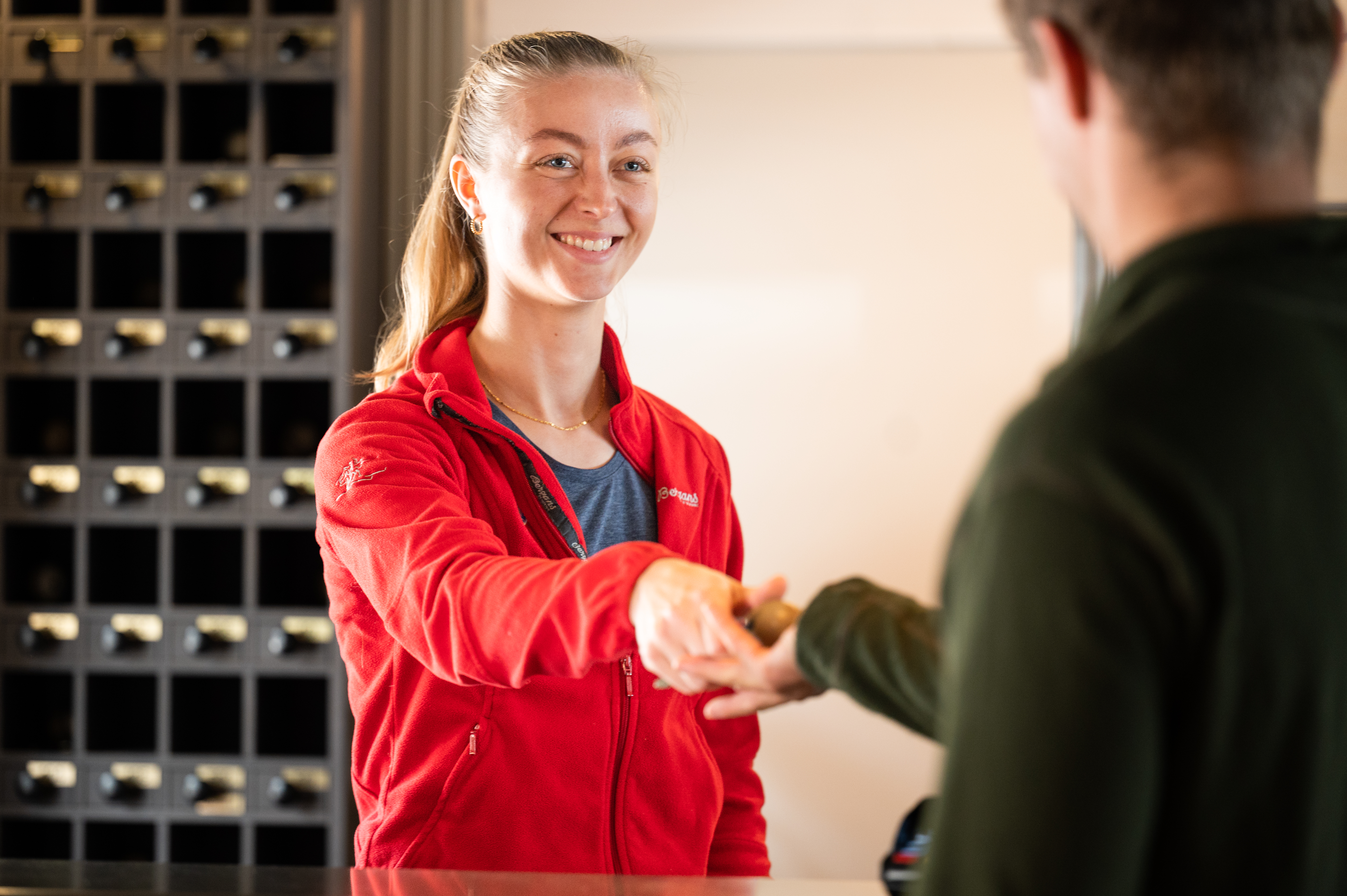 Foto. En jente med r&oslash;d jakke st&aring;r i resepsjonen og gir en romn&oslash;kkel til en gjest.