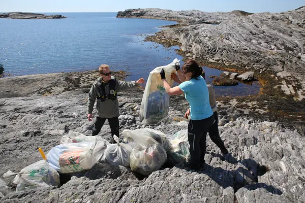 To mennesker rydder plast i strandsona en sommerdag med sol og blå himmel.