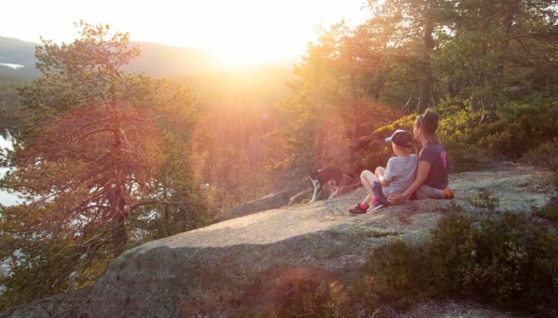 To personer og en hund sitter på et svaberg i skogen og skuer ut over solnedgangen. 