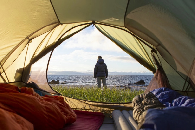 Person står i teltåpningen på stranda og skuer utover havet.