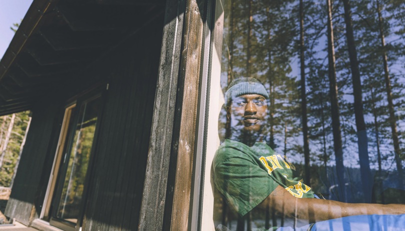 Mann i grønn t-skjorte og blå lue sitter innenfor et vindu og ser ut. I speilbildet i vinduet skimtes høye trær. 