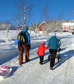 To voksne og et barn triller en barnevogn opp bakken mot en rød gårdsbygning om vinteren, med snødekte bakker rundt. Masse folk er samlet rundt hytta, og noen leker i snøen. 