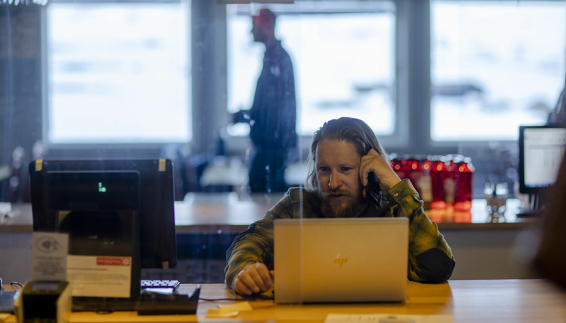 Mann sitter bak en bærbar PC og ser på skjermen mens han holder en mobiltelefon mot øret.