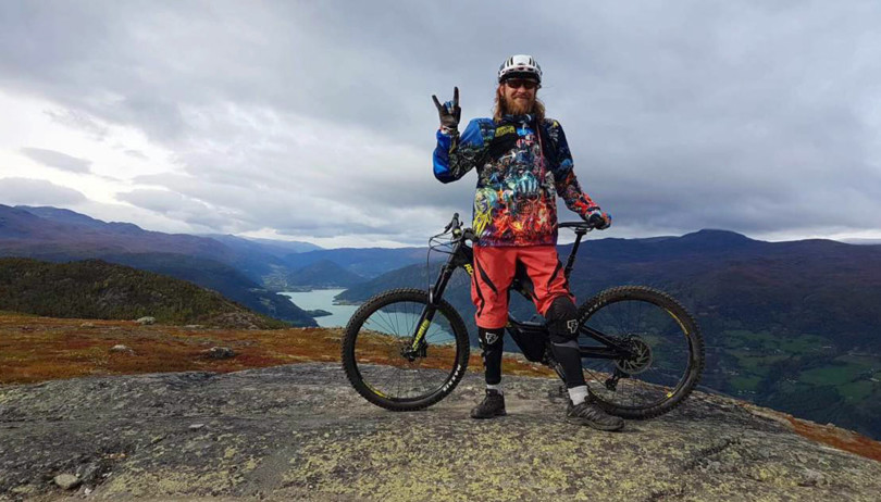 Mann i fargerik jakke og lakserosa shorts lener seg inntil en sykkel på toppen av et fjell.