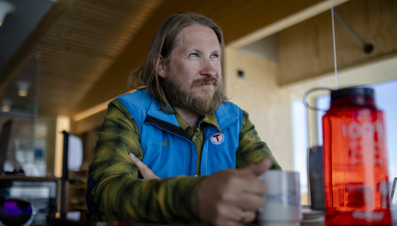 Mann i grønn skjorte og blå vest sitter ved et bord og drikker kaffe mens han ser ut vinduet. 