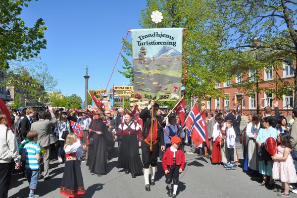 17.-mai tog, hvor vi ser en gruppe kommende mot oss. Foran holder en mann i bunad opp TT sin fane, som står sentralt i bildet. Diverse andre personer i bunad kommer bak ham, og flere barn og voksne står på hver side og ser på. I bakgrunnen kan man se statuen av Olav Tryggvason.