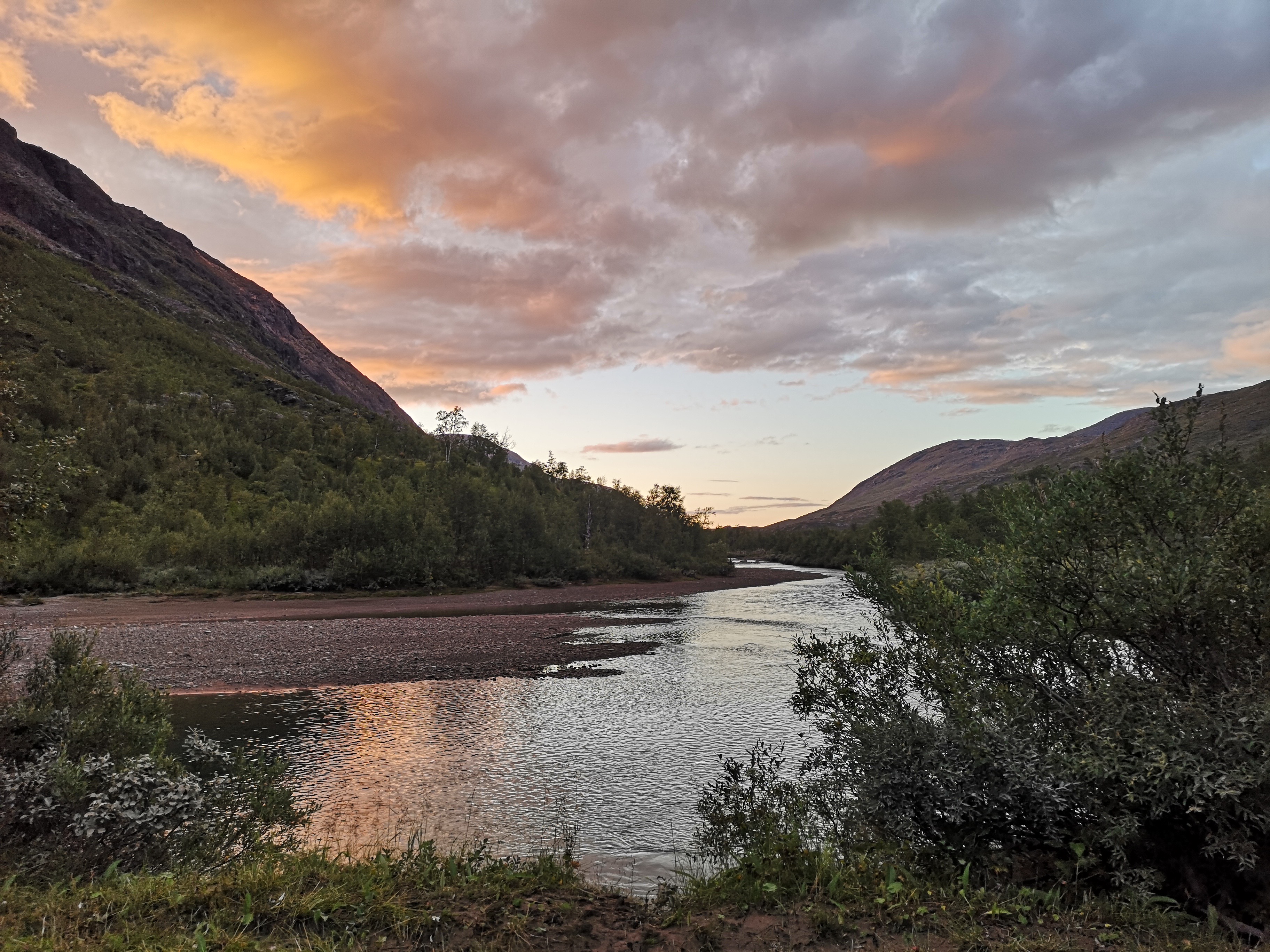 En nydelig kveldshimmel med skyer over en elv. Noe lav vegetasjon i forgrunnen og bjørketrær på andre siden av elva. Høye fjell på begge sider.