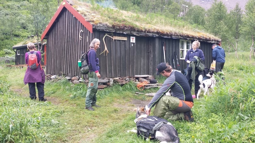Fem mennesker i turklær og to hunder med kløv står foran ei mørkebrun hytte med torvtak. Det er lauvtrær bak hytta.