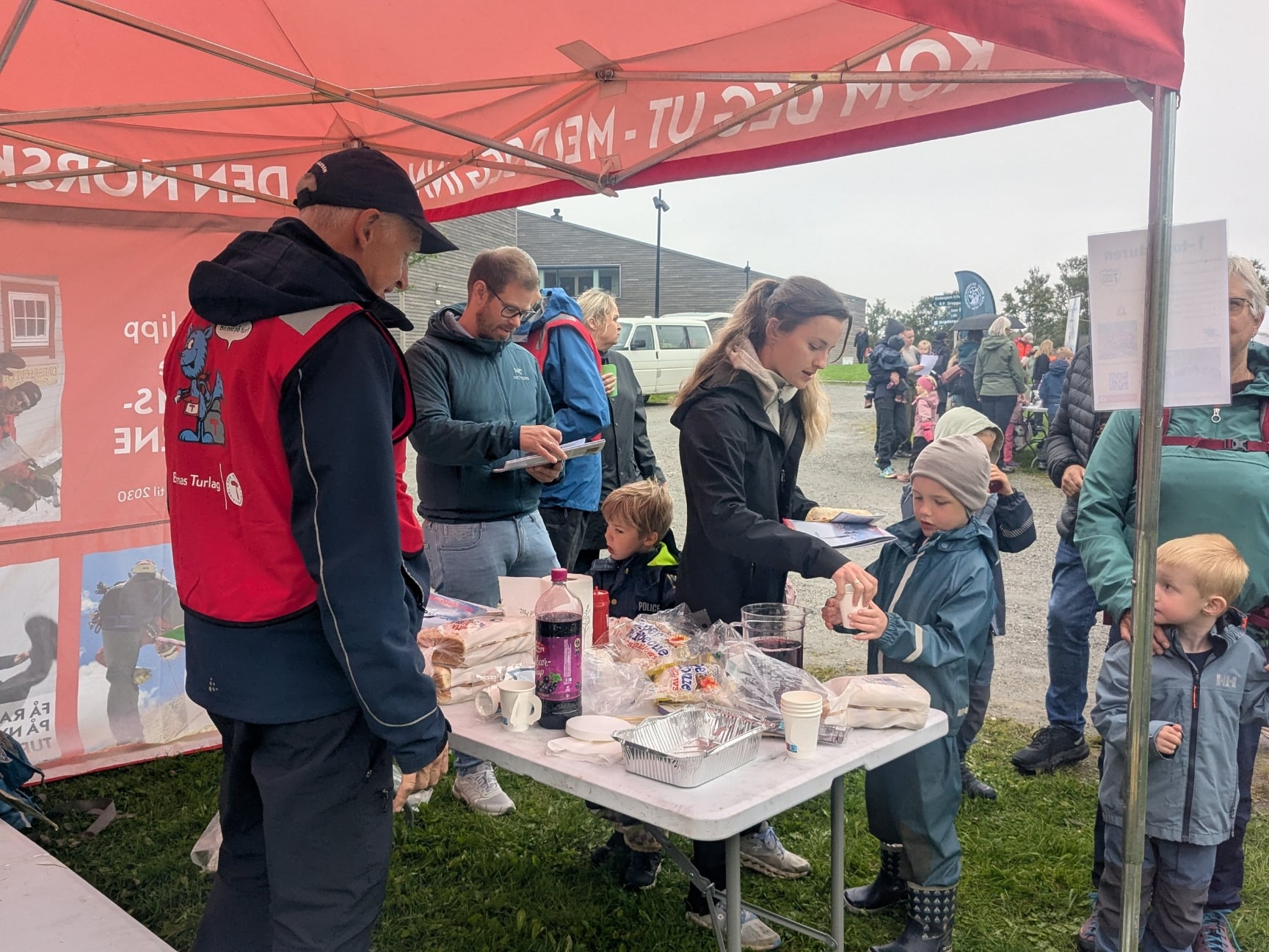 Voksne og barn foran standbord under taket til r&oslash;d paviljong