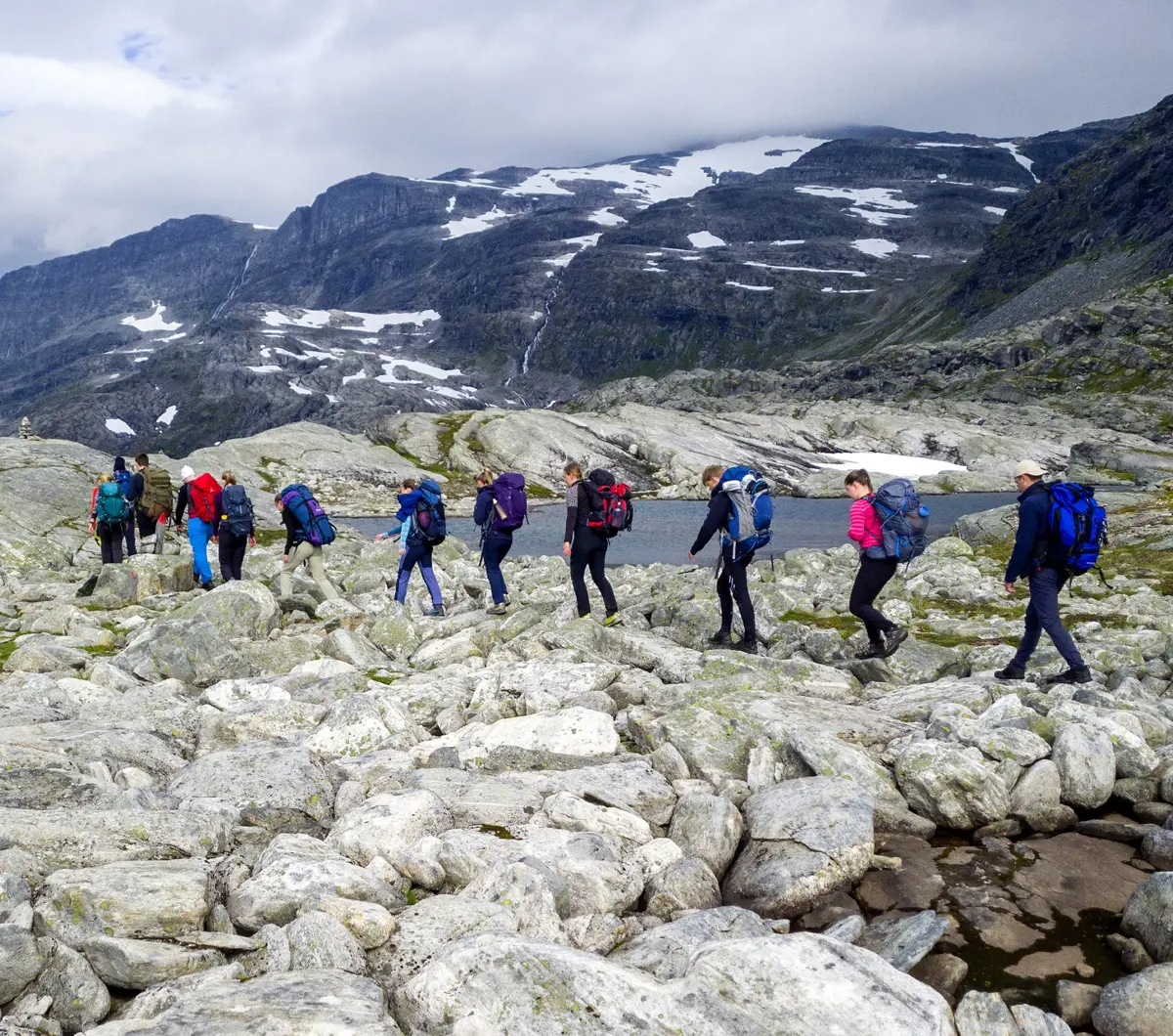 Skoleklasse med ungdom går til fots blant høye fjell i steinete terreng