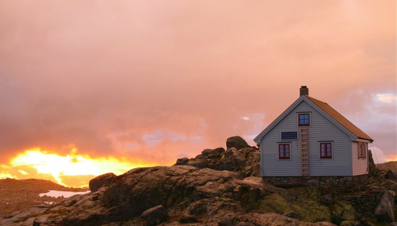 Grå hytte på toppen av en fjellknaus. Sola skinner gjennom et grått skydekke og farger hytta og naturen rød.
