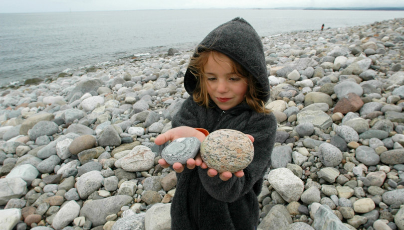 Barn med grå hettejakke står på en strand med runde steiner og holder opp en stein i hver hånd.