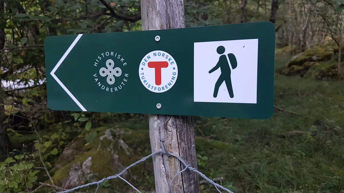 Grønt retningsskilt med logo historisk vandrerute, dnt sin raude T og person med sekk