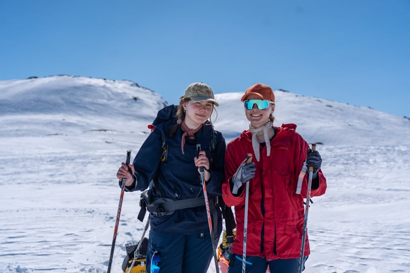 To blide jenter som smiler til kamera. De holder skistaver i hendene.