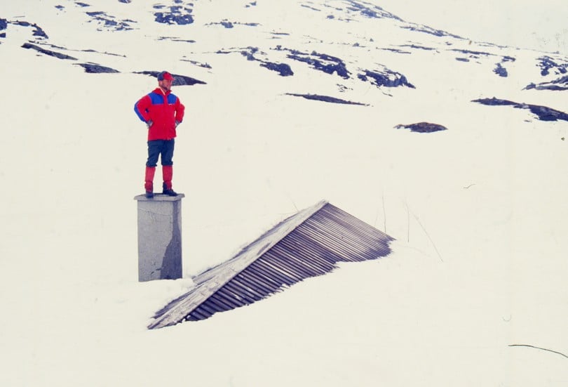 En mann med rød jakke som står oppå en pipe på en hytte som er nedsnødd.