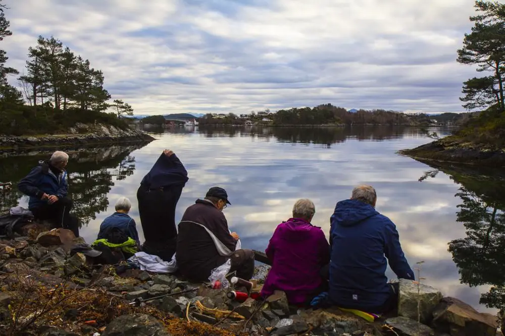 Rastepause for seniorar i strandkanten med skue utover ein speilblank fjord