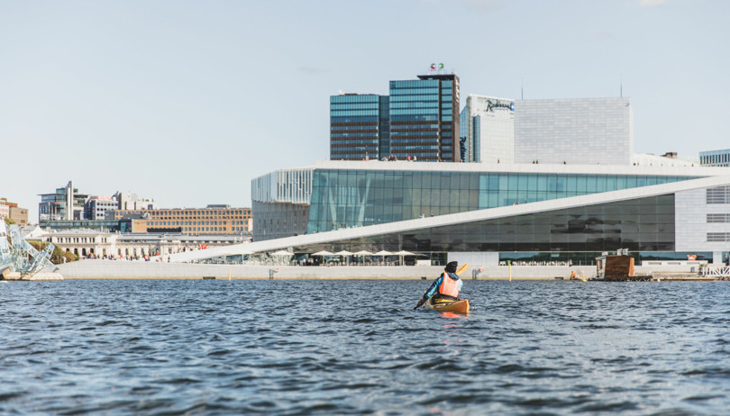 Person i gul kajakk padler innover mot by med høye bygninger.