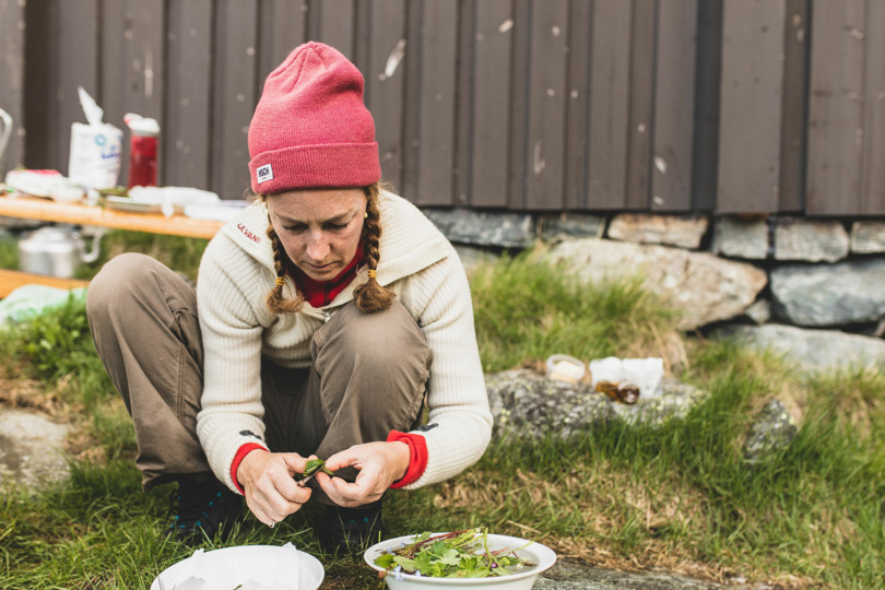 Kvinne med rosa lue sitter på huk på bakken og lager mat foran en hytte.